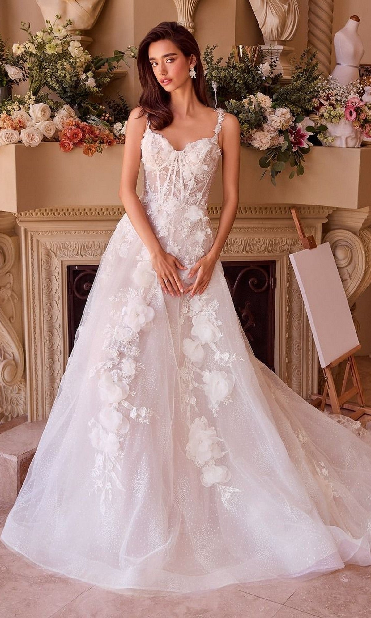 A woman in the Andrea and Leo WL045 long wedding dress with a lace corset bodice stands indoors before a flower-adorned mantle and an empty easel.